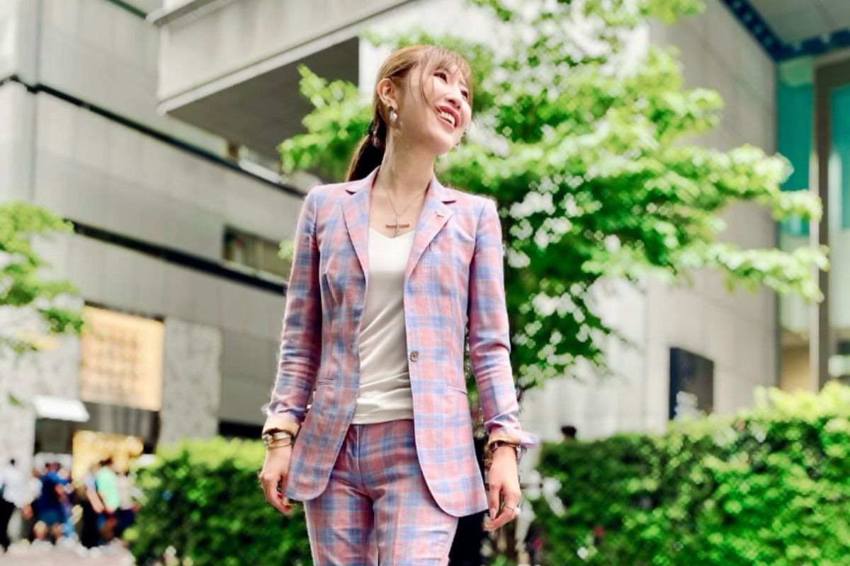 ピンクとブルーのチェック柄スーツを着た勝友美さんが、緑の街路樹を背に屋外で笑顔で立つ全身ショット