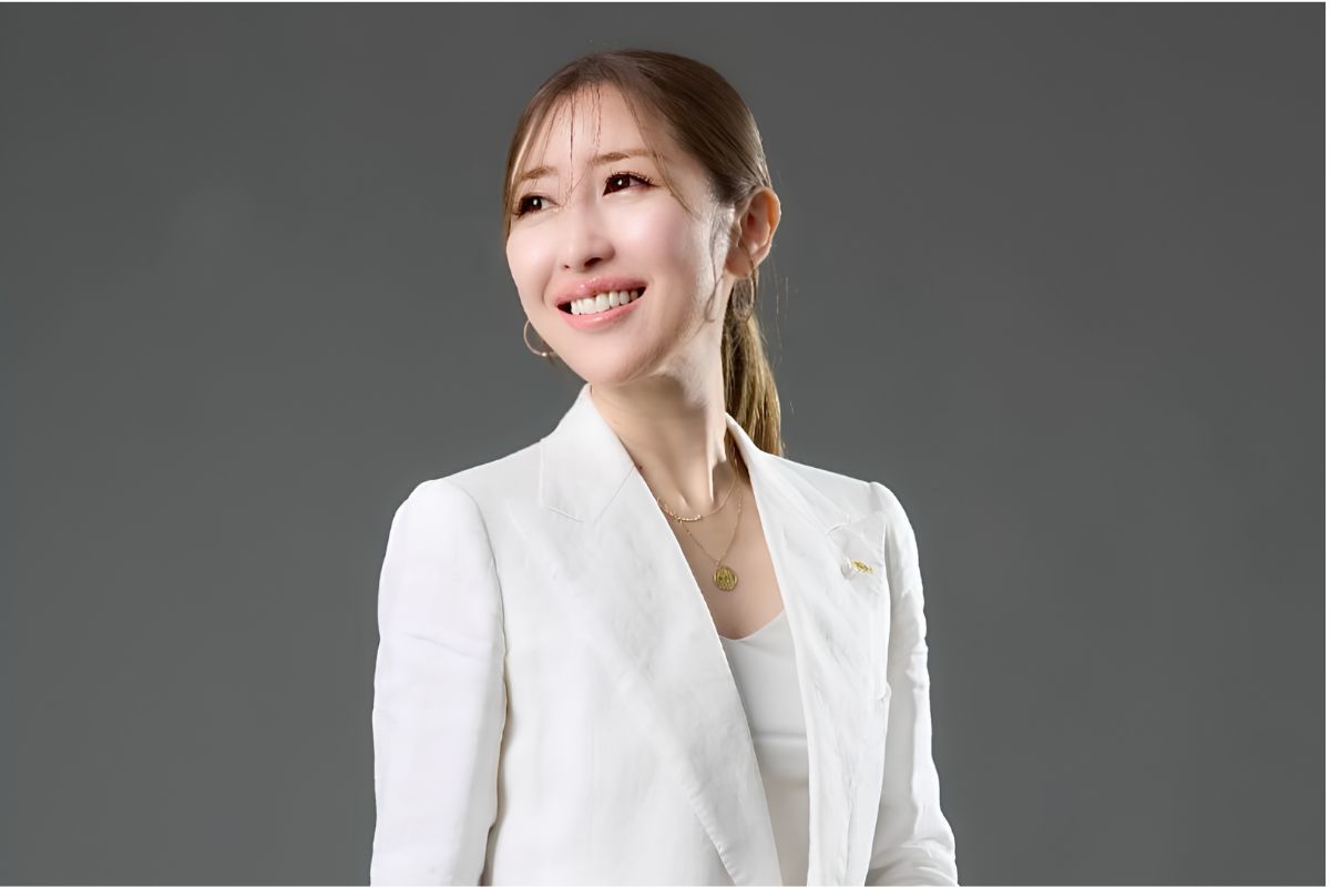 オーダースーツブランドRe.museの代表でテーラーの勝友美さんが、白のリネンセットアップ姿でグレー背景の前に立ち笑顔を見せるポートレート
